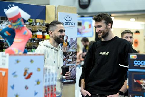 Two men chat and smile while standing near a booth of BV Sport at IRX